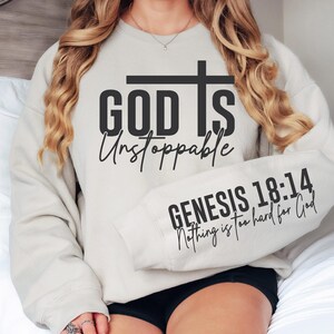 May include: A light beige sweatshirt with the words "GOD IS Unstoppable" and a cross graphic in black. The sleeve displays "GENESIS 18:14 Nothing is too hard for God" in black script. The sweatshirt is worn by a person.