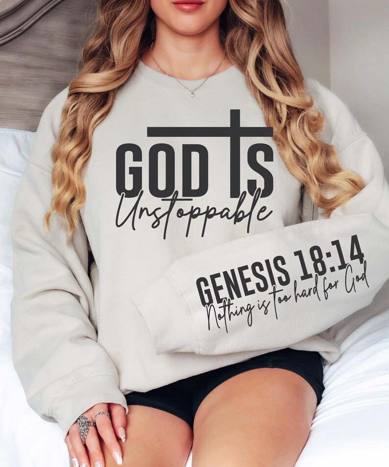 May include: A light beige sweatshirt with the words "GOD IS Unstoppable" and a cross graphic in black. The sleeve displays "GENESIS 18:14 Nothing is too hard for God" in black script. The sweatshirt is worn by a person.