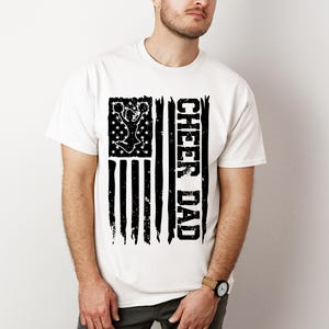 May include: White t-shirt featuring a black and white distressed American flag design. The flag depicts a cheerleader silhouette and the text "Cheer Dad".