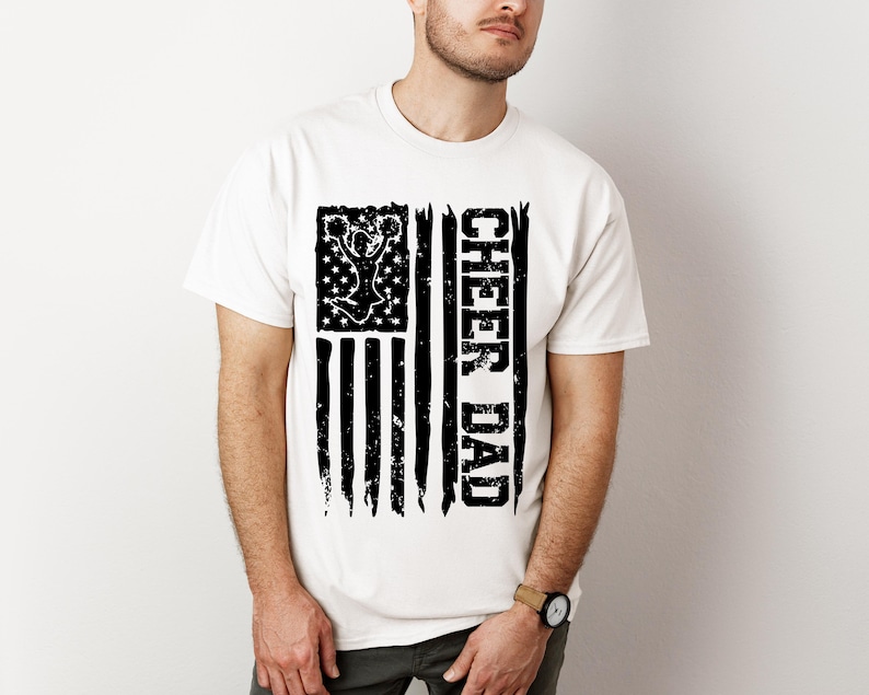 May include: White t-shirt featuring a black and white distressed American flag design. The flag depicts a cheerleader silhouette and the text "Cheer Dad".