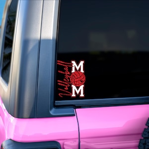 Peut inclure: Un Jeep rose avec une vitre noire orn&eacute;e d'un autocollant rouge paillet&eacute; "Volleyball Mom". L'autocollant comprend un graphisme de ballon de volley-ball entre les mots "MOM". L'arri&egrave;re-plan montre une sc&egrave;ne de plage avec des palmiers.