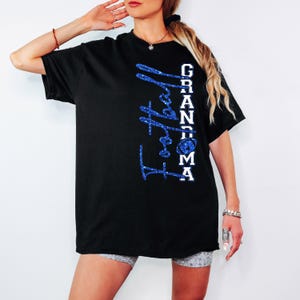 Pu&ograve; includere: T-shirt nera con la scritta "Football Grandma" in glitter blu. La parola "Grandma" &egrave; in lettere bianche. La maglietta presenta una grafica di football americano.