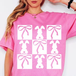 Op de afbeelding: Roze T-shirt met een wit grafisch ontwerp van negen vierkanten. Elk vierkant heeft een wit konijn met een strik.