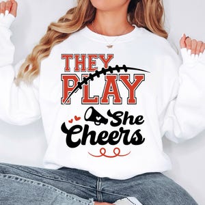 May include: White sweatshirt with the text "They Play She Cheers" in black and red lettering. A football and a cheerleader's megaphone are included in the design.