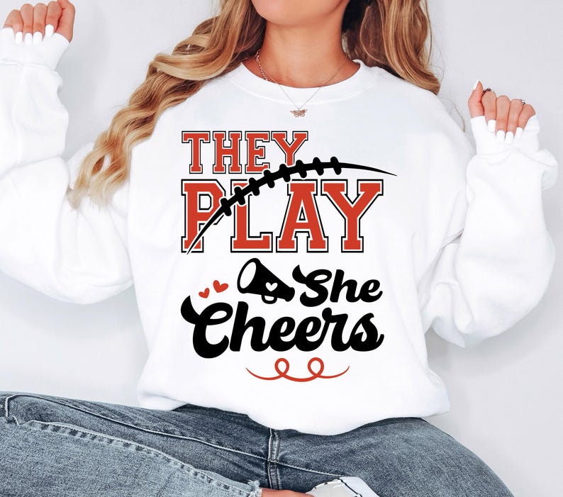 May include: White sweatshirt with the text "They Play She Cheers" in black and red lettering. A football and a cheerleader's megaphone are included in the design.