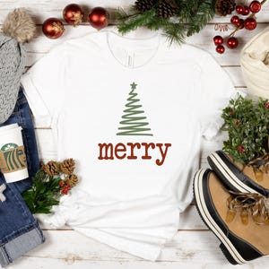 May include: A white t-shirt with a green Christmas tree design and the word "merry" in red. The image includes a gray beanie, blue jeans, a coffee cup, and tan winter boots.
