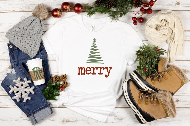 May include: A white t-shirt with a green Christmas tree design and the word "merry" in red. The image includes a gray beanie, blue jeans, a coffee cup, and tan winter boots.