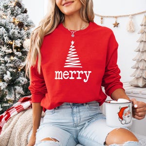 May include: A red sweatshirt with a white Christmas tree design and the word "merry" in white. The person is holding a mug with a Santa Claus illustration. The background includes a decorated Christmas tree.