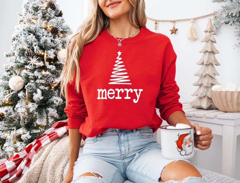 May include: A red sweatshirt with a white Christmas tree design and the word "merry" in white. The person is holding a mug with a Santa Claus illustration. The background includes a decorated Christmas tree.
