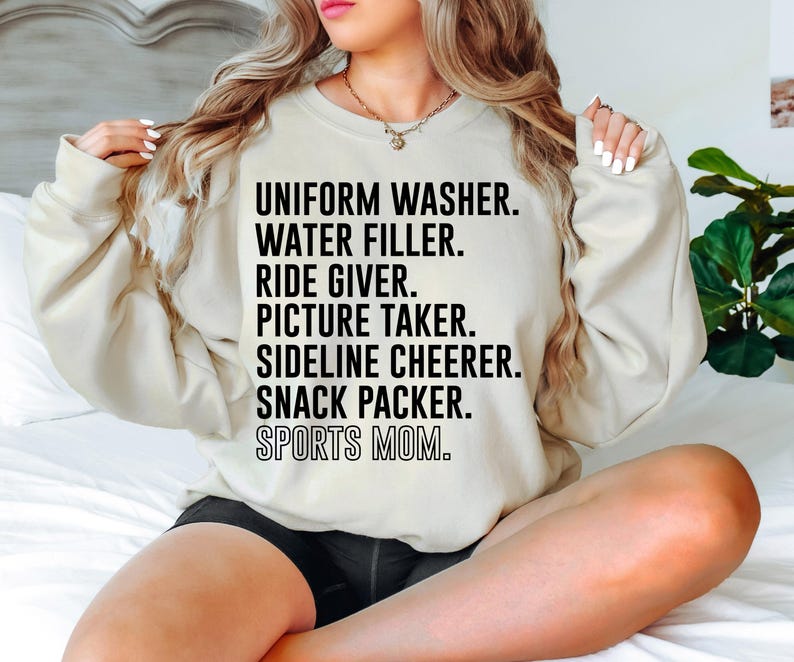 May include: A cream-colored sweatshirt with black text that reads "UNIFORM WASHER. WATER FILLER. RIDE GIVER. PICTURE TAKER. SIDELINE CHEERER. SNACK PACKER. SPORTS MOM."