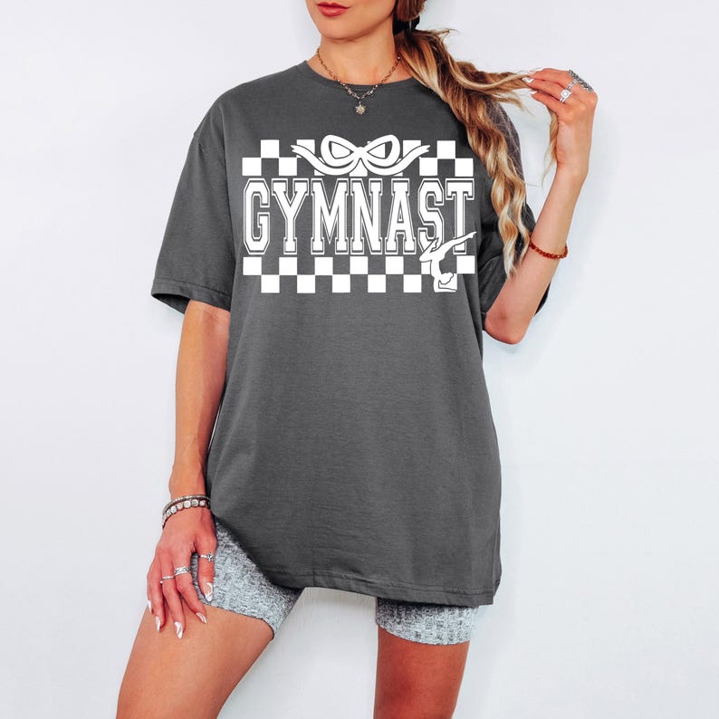 May include: A dark gray oversized t-shirt with a white checkered pattern and the text "GYMNAST" in white. The text is in a bold, blocky font. A white silhouette of a gymnast performing a handstand is to the right of the text.