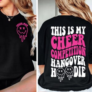 May include: Black sweatshirt with the text "CHEER MOM" and a pink smiley face graphic on the front. The back of the shirt features the text "THIS IS MY CHEER COMPETITION HANGOVER HOODIE" in white and pink.