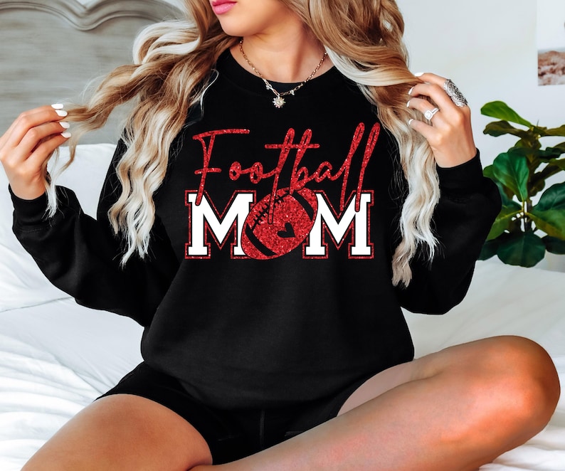 May include: Black sweatshirt with the text "Football MOM" in red glitter lettering and a red football graphic. The word "MOM" is in white block letters with a red outline. A heart is in the center of the football.