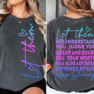 May include: Dark gray t-shirt with the words "Let them" in a gradient of blue and pink. The back of the shirt features the text "Misunderstand You, Judge You, Gossip and Doubt You. Your Worth Has Already Been Affirmed By God. Keep Shining."