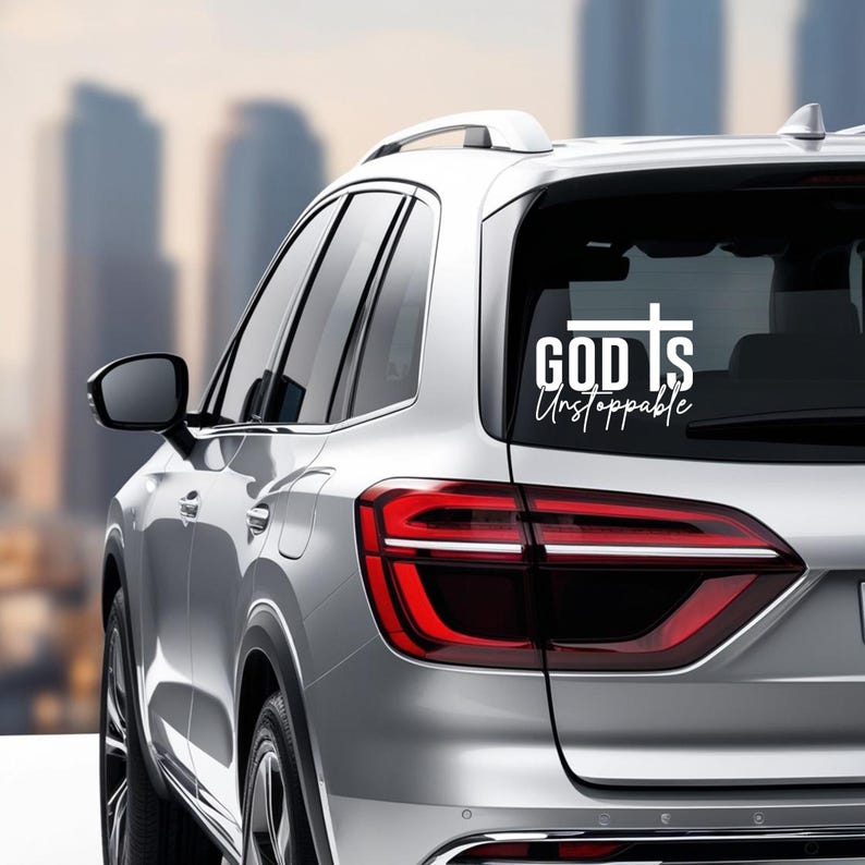 May include: A white decal on the rear window of a silver car. The decal features the words "GOD Is Unstoppable" in a mix of bold and script fonts, with a cross symbol above the word "Is". The car is parked in front of a blurred cityscape.