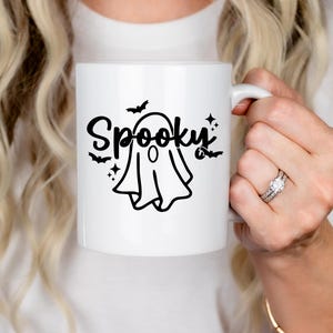Op de afbeelding: Een witte keramische koffiemok met een zwarte geest en de tekst "Spooky" in een zwart cursief schrift. De geest heeft een zwarte omtrek en een zwarte cirkel voor een oog.