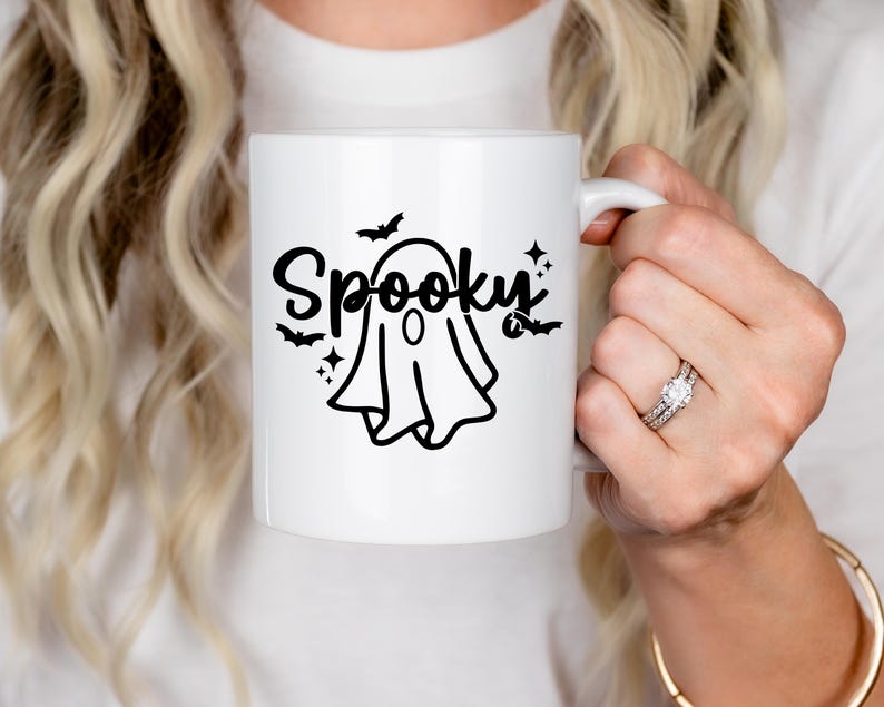 Op de afbeelding: Een witte keramische koffiemok met een zwarte geest en de tekst "Spooky" in een zwart cursief schrift. De geest heeft een zwarte omtrek en een zwarte cirkel voor een oog.