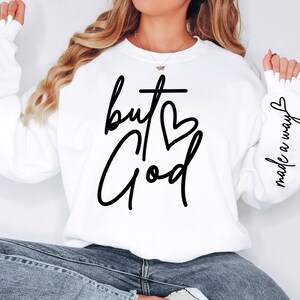 May include: White sweatshirt with the text "but God" in black script with a cross and heart. The right sleeve has the text "made a way" in black script.