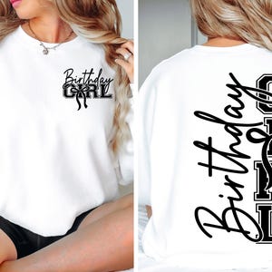 May include: White sweatshirt featuring the text "Birthday Girl" in black script and block letters, with a bow design. The text is printed on both the front and back of the garment.