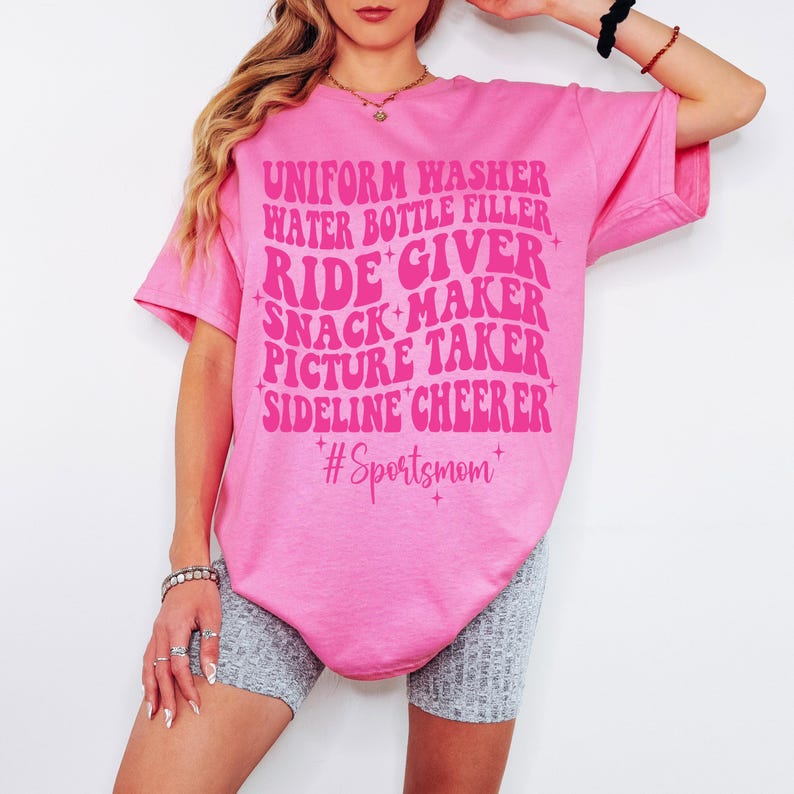 May include: Pink t-shirt with a retro font design that reads "Uniform Washer, Water Bottle Filler, Ride Giver, Snack Maker, Picture Taker, Sideline Cheerer" and the hashtag "#Sportsmom".