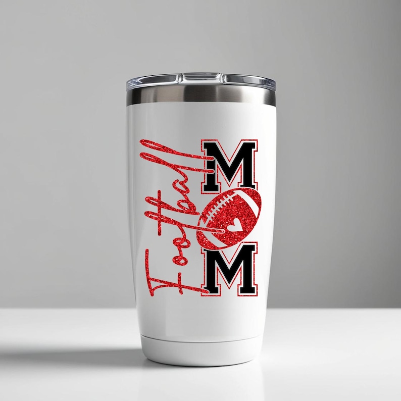 May include: White tumbler with a silver lid featuring the words "Football Mom" in red glitter script. The design includes a red football with a heart and the letters "MOM" in black.