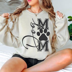 Puede incluir: Una sudadera de color crema con una huella de pata negra y el texto "Mom" en letras negras.