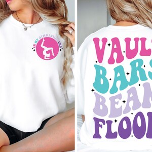 May include: White sweatshirt with a pink circle on the front that says "In My Gymnastic Era" with a silhouette of a gymnast in the circle. The back of the sweatshirt has the words "Vault Bars Beam Floor" in a retro font with pink, blue, and purple colors.