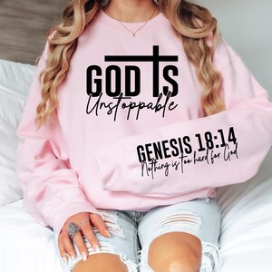May include: A light pink sweatshirt with the words "GOD Is Unstoppable" and a cross graphic in black. The sleeve displays the text "GENESIS 18:14 Nothing is too hard for God" in black. The sweatshirt is a crew neck style.