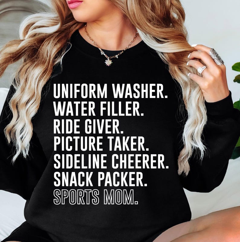 May include: Black sweatshirt with white text that reads "UNIFORM WASHER. WATER FILLER. RIDE GIVER. PICTURE TAKER. SIDELINE CHEERER. SNACK PACKER. SPORTS MOM."
