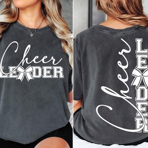 Puede incluir: Una camiseta gris oscuro con el texto "Cheer Leader" impreso en la parte delantera y el texto "Cheer Leader" impreso verticalmente en la parte posterior. El texto es blanco e incluye un lazo blanco.