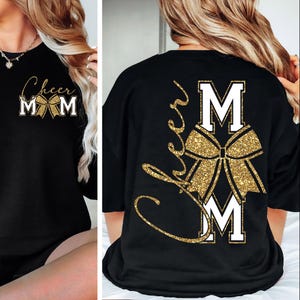 Peut inclure: T-shirt noir à manches longues avec l'inscription "Cheer Mom" en lettres dorées et un motif de nœud pailleté doré. Le t-shirt comporte également les lettres "MOM" en lettres blanches.