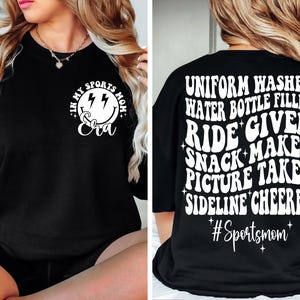 May include: Black t-shirt with white text that reads "In my sports mom era" with a smiley face and lightning bolts on the front. The back of the shirt reads "Uniform washer, water bottle filler, ride giver, snack maker, picture taker, sideline cheerleader #Sportsmom".