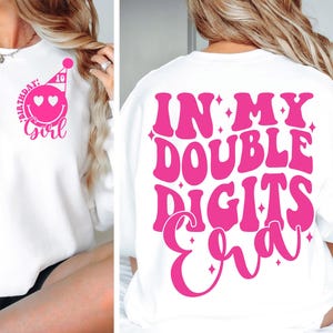 Könnte beinhalten: Weißer Pullover mit rosa Schriftzug "In My Double Digits Era" auf der Rückseite. Die Vorderseite des Pullovers hat ein rosa Smiley mit einer Partymütze und den Text "Birthday Girl" mit der Zahl 10 über dem Smiley.