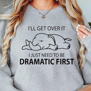 May include: A pale grey sweatshirt featuring the text "I'll get over it, I just need to be dramatic first" and a black illustration of a sleeping elephant. The sweatshirt has long sleeves and a crew neck.