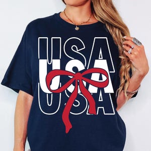 Op de afbeelding: Marineblauwe t-shirt met het woord "USA" in witte blokletters en een rode strik. Een casual top, geschikt voor patriottische evenementen of dagelijks gebruik.