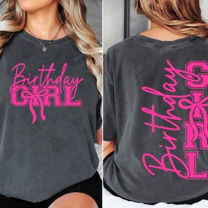May include: Charcoal grey t-shirt with the words "Birthday Girl" in pink script and block letters, featuring a bow graphic. The same design is on the back, vertically. A casual, comfortable tee for a birthday celebration.