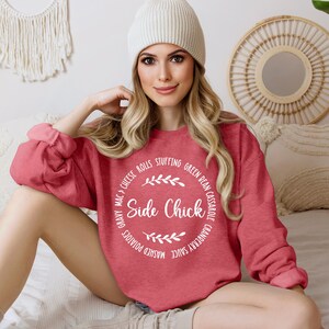 May include: A red sweatshirt with a white circle graphic that says "Side Chick" and lists various Thanksgiving foods around the circle. The model is wearing a white beanie.