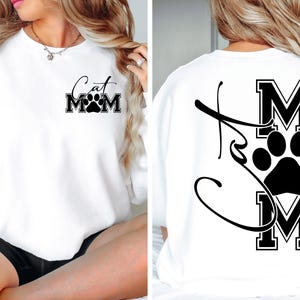 May include: White sweatshirt with the text "Cat Mom" and a paw print design. The text is in a black, stylized font. The back of the sweatshirt also features a paw print and the letters "MOM" in a black, block font.