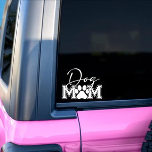 Puede incluir: Un coche rosa con una calcoman&iacute;a de vinilo blanco que dice "Dog Mom" con una huella de pata en el medio de la palabra "Mom".