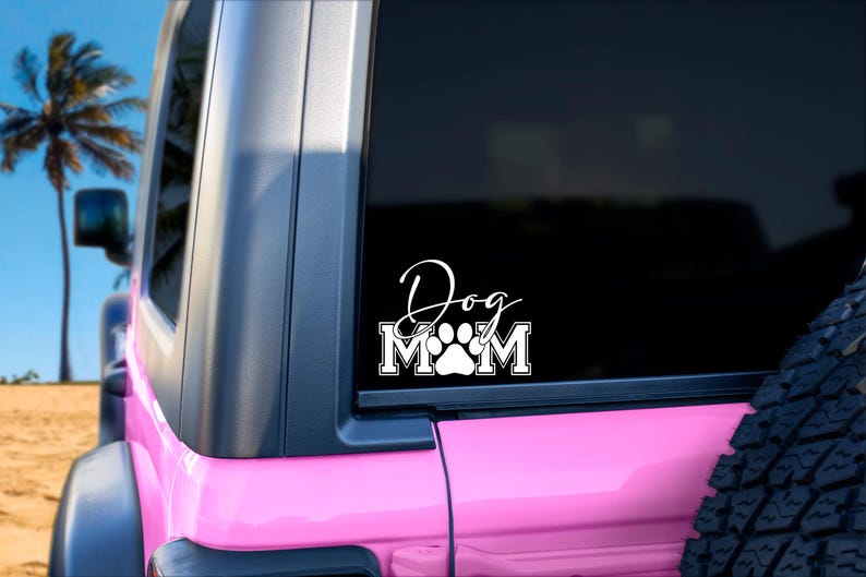 Puede incluir: Un coche rosa con una calcoman&iacute;a de vinilo blanco que dice "Dog Mom" con una huella de pata en el medio de la palabra "Mom".