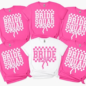 May include: Five t-shirts with the text "Bride Squad" in white letters on a pink and white checkered background. The shirts are folded and arranged in a circle.