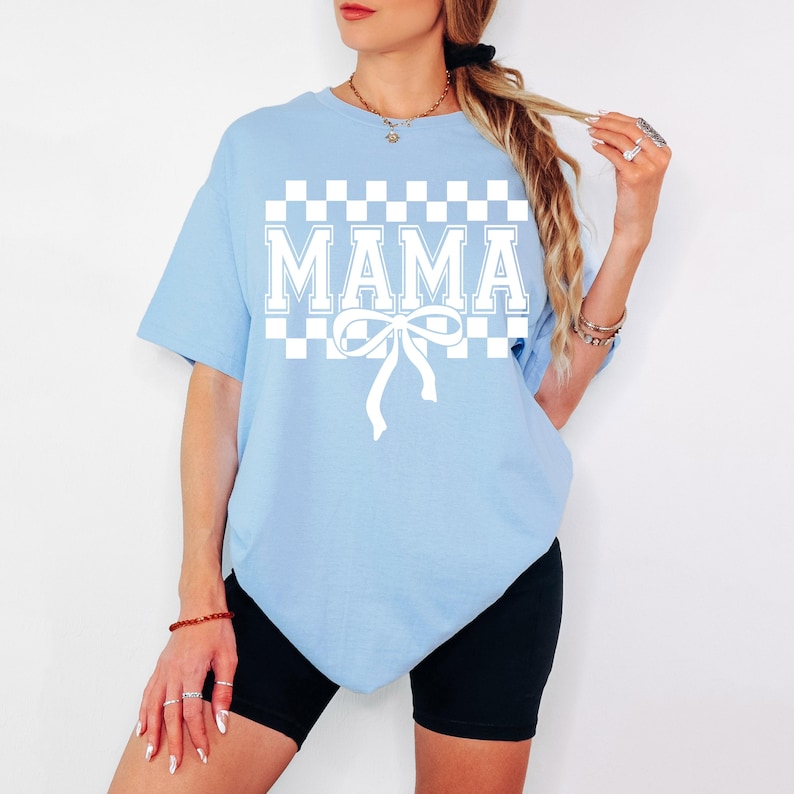 May include: A light blue t-shirt with a white checkered pattern and the word "MAMA" in white letters. A white bow is centered below the text.
