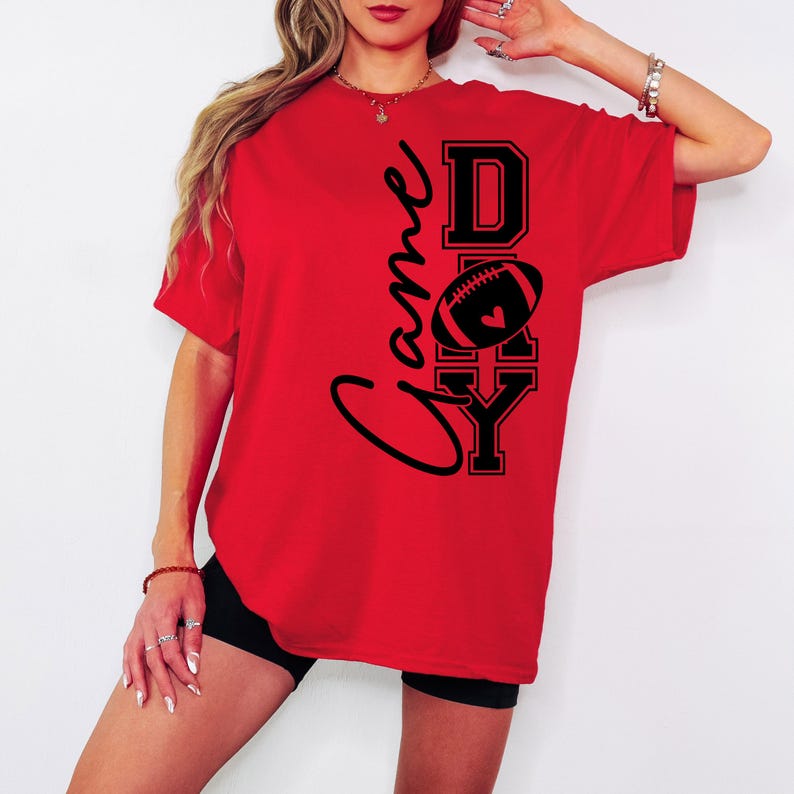 May include: Red t-shirt with the words "Game Day" in black script and block letters. A football with a heart is in the center. The shirt is a casual, oversized style.
