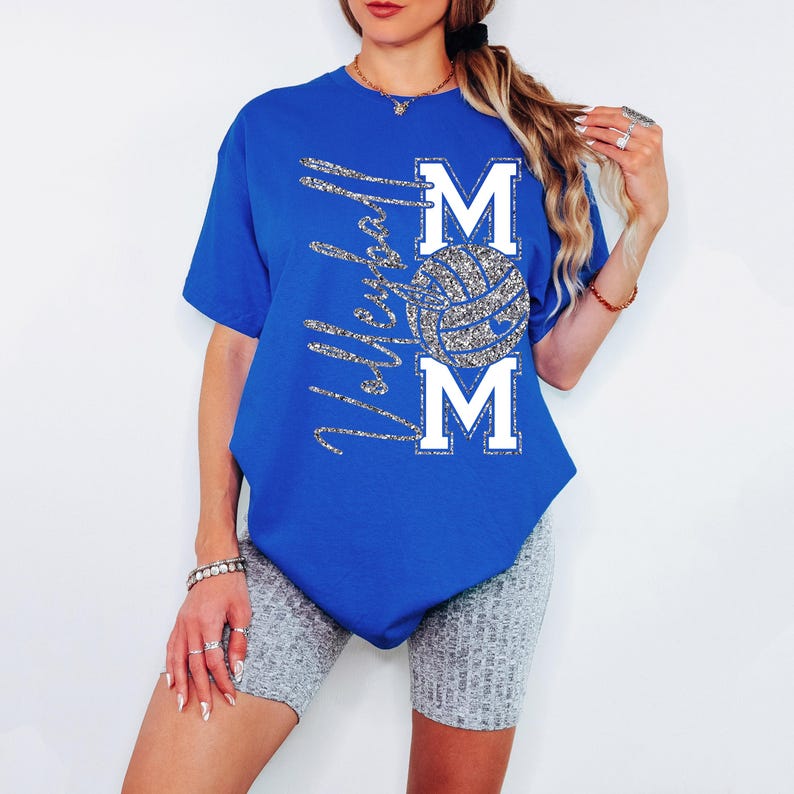 May include: Royal blue t-shirt with the text "Volleyball Mom" in silver glitter script. The word "MOM" is in white block letters, with a glitter volleyball in the center. The shirt is a short-sleeved crew neck.