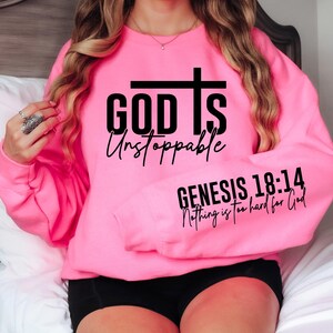 May include: A bright pink sweatshirt with the words "GOD IS Unstoppable" and a cross graphic. The sleeve displays the text "GENESIS 18:14 Nothing is too hard for God" in black lettering. The sweatshirt is a crew neck style.