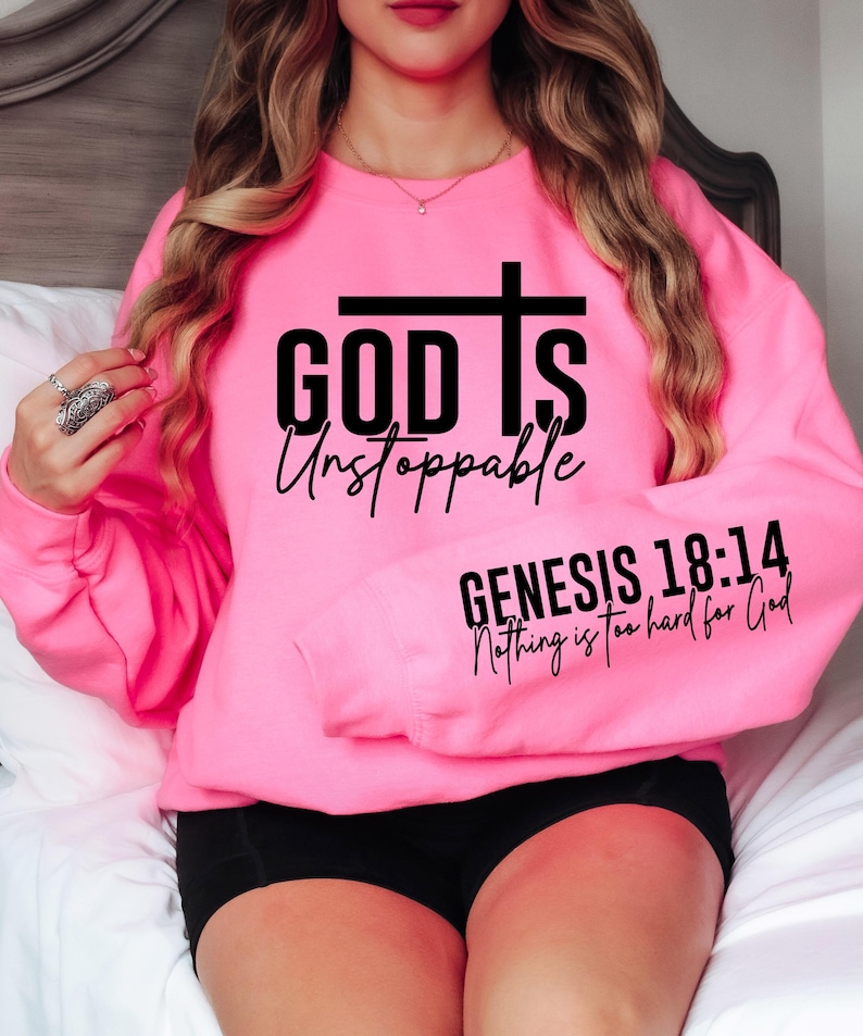 May include: A bright pink sweatshirt with the words "GOD IS Unstoppable" and a cross graphic. The sleeve displays the text "GENESIS 18:14 Nothing is too hard for God" in black lettering. The sweatshirt is a crew neck style.