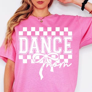 以下が含まれることがあります： 白のチェック柄のピンクのTシャツ。白で「DANCE MOM」と書いてあり、白いリボンがついています。
