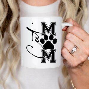 May include: White ceramic mug with the word "Cat Mom" in a script font, with the letters "MOM" in a bold, block letter style, and a black paw print in the center. The mug has a white handle.