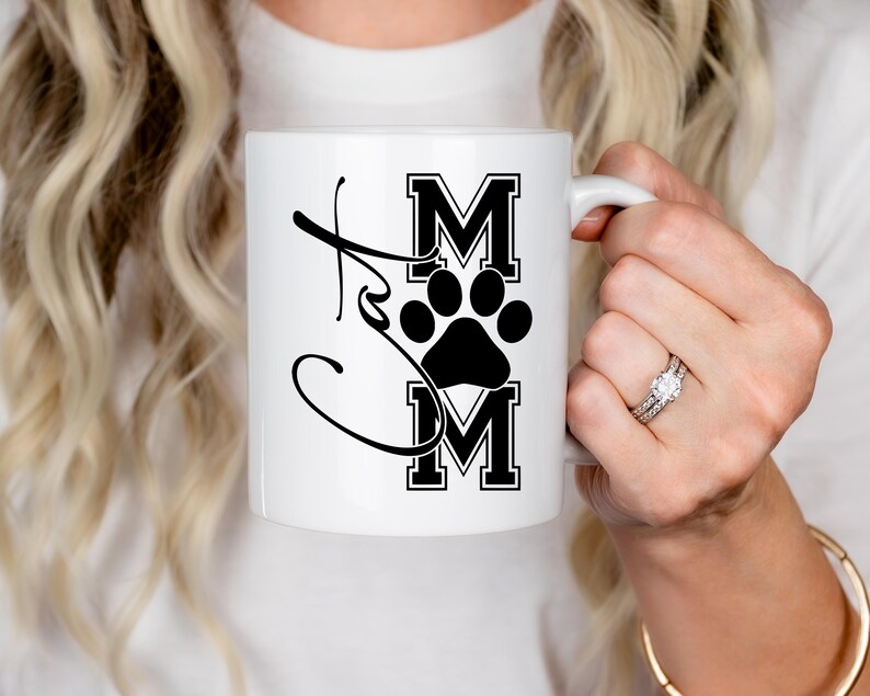 May include: White ceramic mug with the word "Cat Mom" in a script font, with the letters "MOM" in a bold, block letter style, and a black paw print in the center. The mug has a white handle.