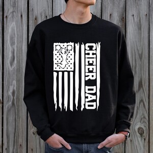May include: Black sweatshirt with a white distressed American flag design. The flag features a cheerleader silhouette and the text "Cheer Dad".